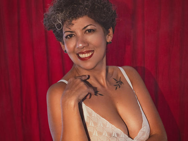 JuliaNewell, a curvy woman with short curly hair, wears a white lace bodysuit, exposing her tits and pussy, standing with a hand on her head, smiling.