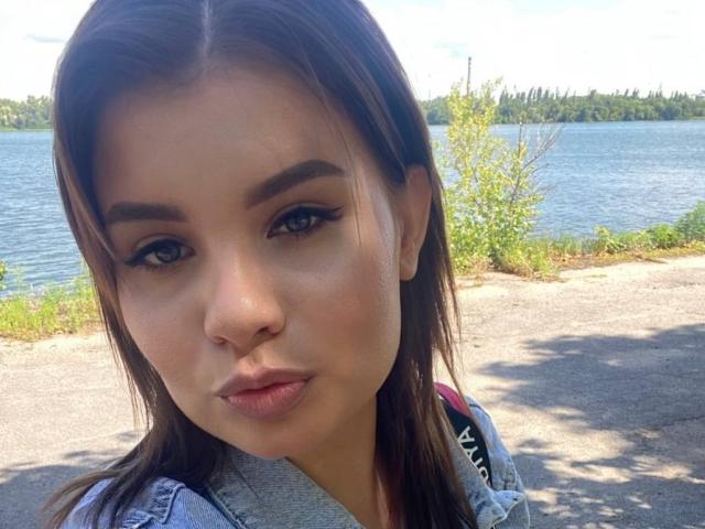 A close-up photo of AmandaHoppe with light skin and brown hair. She has blue eyes, dark eyebrows, and wears black eyeliner. Her expression is neutral with slightly parted lips. She's wearing a denim jacket. The background shows a blue lake with green trees. The image is sharply focused on AmandaHoppe's face. Natural light highlights her features. The composition centers AmandaHoppe in the frame.
