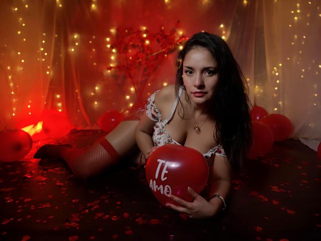 AngelaBranson lies on a dark floor, surrounded by red balloons and petals. She has long, dark hair, and wears a floral off-shoulder top with red fishnet stockings. She holds a red balloon with "TE AMO" written on it. The background is illuminated by warm, yellow fairy lights. AngelaBranson's intense gaze is directed at the camera. Her skin is fair, and she has a necklace with a small pendant. The image has a red and yellow color palette, with a sensual and romantic vibe. The composition is centered on AngelaBranson and the red balloon.