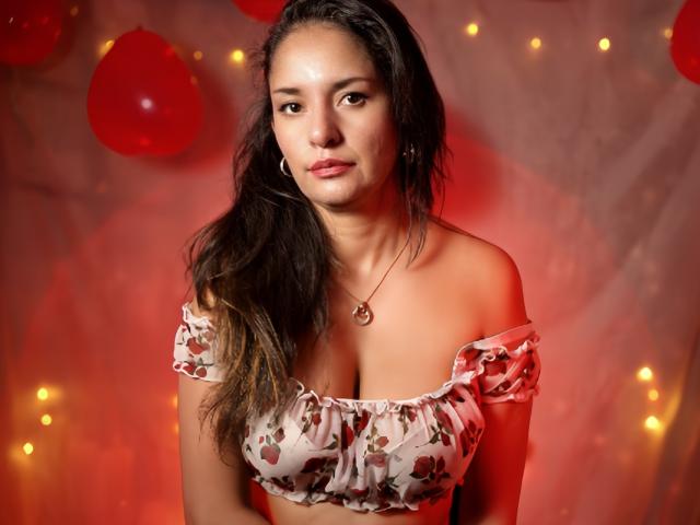AngelaBranson with wavy brown hair, off-shoulder floral top, medium tits, red necklace, soft red lighting, looking at camera, slight smile, red balloons in background.