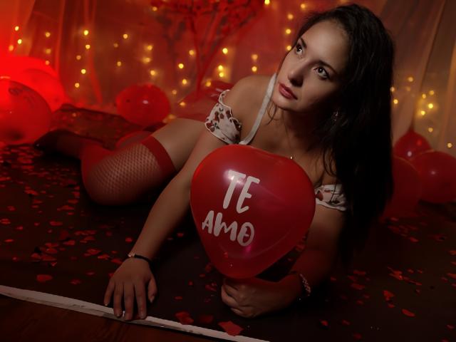 AngelaBranson is lying on a dark floor, holding a red heart-shaped balloon with "TE AMO" written in white. She has dark, wavy hair and is wearing a white, off-shoulder top with red heart patterns and red fishnet stockings. Her eyes are looking directly at the camera. Red balloons and petals are scattered around her, with soft, warm yellow lights in the background. Her expression is serious and slightly sultry. The image is highly saturated with red and yellow tones.