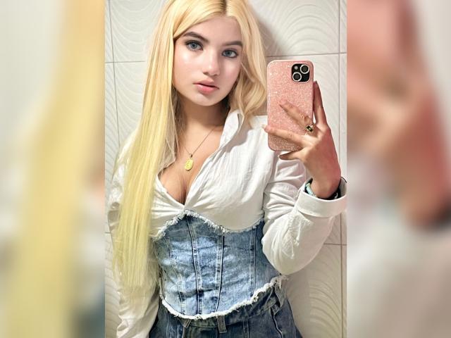 SabrinaColle mit langen blonden Haaren, weißem Hemd, Jeans-Korsett, hält ein rosa Handy, macht ein Selfie im Spiegel, zeigt kleine Brüste, dekolletiert, blickt mit leicht geöffneten Lippen in die Kamera..
