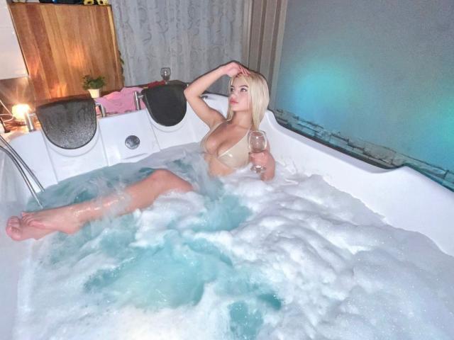 SabrinaColle, blonde, in a bubble-filled hot tub, holding a glass, with her tits exposed, her ass partially visible, one leg up, and a hand on her head.