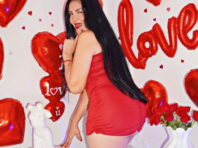 AlizonMoreti with long black hair, red dress, and red lipstick, smiling, showing her ass, surrounded by red heart balloons and roses. Her right hand touches her chin.