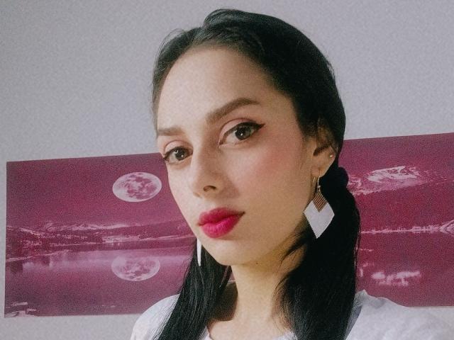AnaisHilton has black pigtails, pink lipstick, and white diamond earrings. She's looking at the camera with brown eyes, wearing a white top. Her tits are not visible.