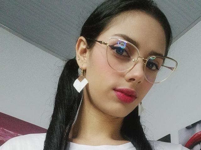 AnaisHilton has dark pigtails, pink lipstick, and wears gold-rimmed glasses with white earrings. Her blue eyes reflect a light source. She's wearing a white shirt.