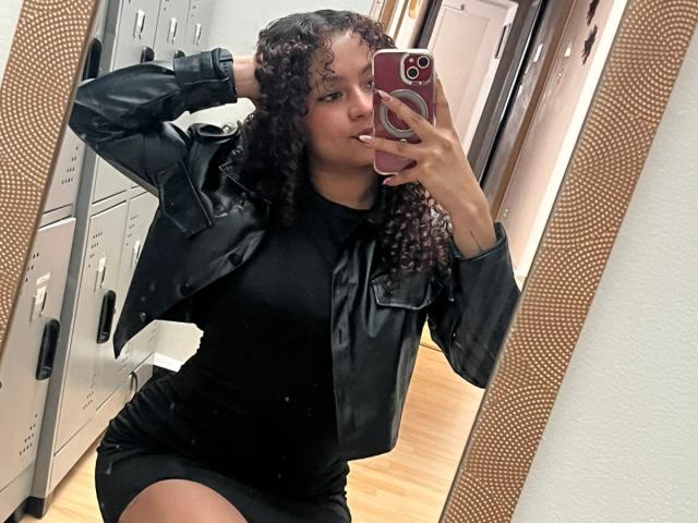 SofiaPassionne with curly hair, wearing a black leather jacket and tight black dress, posing in front of a mirror, holding a pink phone, showing her ass.