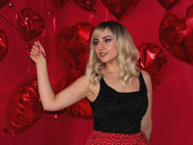 MilenaBaldwin with blonde wavy hair, black lace top, red polka dot skirt, red heart balloons, smiling, right hand up, red background, cute, sexy, small tits, medium ass.