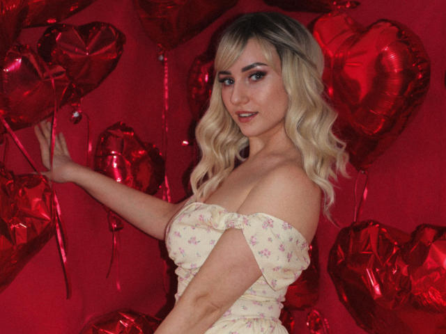 MilenaBaldwin with blonde curls, wearing a floral off-shoulder top, holding red heart balloons, showing her tits, smiling at the camera, red background.