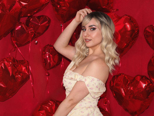 MilenaBaldwin with blonde wavy hair, wearing an off-shoulder floral dress, standing against red heart-shaped balloons, hand in hair, smiling, showing her tits, cute, sexy, red background.