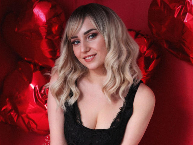 MilenaBaldwin with blonde wavy hair, black lace dress, and cleavage, smiling at the camera, red heart balloons in the background, cute and sexy.