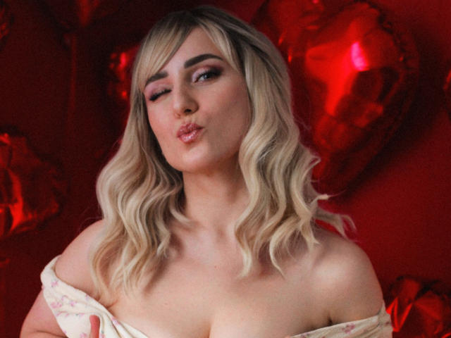 MilenaBaldwin with blonde wavy hair, pink eyeshadow, and pouty lips, off-shoulder floral top, showing her tits, red heart balloons in background, winking, soft lighting.