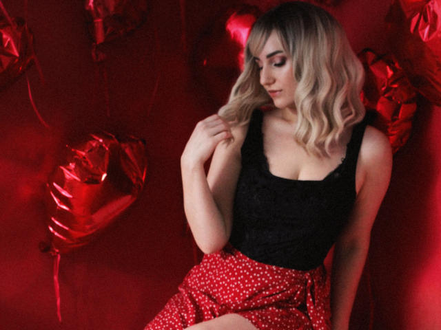 MilenaBaldwin with blonde wavy hair, black lace top, and red polka-dot skirt, touching her hair, red heart balloons in the background, red light, cute, attractive.
