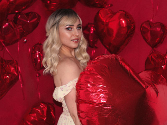 MilenaBaldwin with blonde wavy hair, wearing an off-shoulder floral dress, holding a red heart-shaped balloon, surrounded by red heart balloons, smiling with subtle makeup.