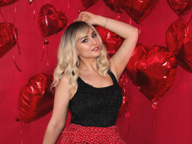 MilenaBaldwin with blonde hair, wearing a black lace top and red polka-dot skirt, posing with arm raised, surrounded by red heart-shaped balloons, showing her tits and ass.
