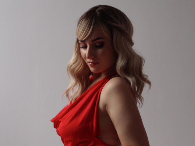 MilenaBaldwin with wavy blonde hair, wearing a red dress, looking down, showing her back and side, with her ass slightly visible, in a soft, indoor light.
