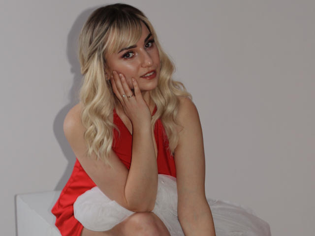 MilenaBaldwin with blonde wavy hair, red sleeveless top, and white skirt, resting her chin on her hand, smiling softly, showing her ring and cute ass.