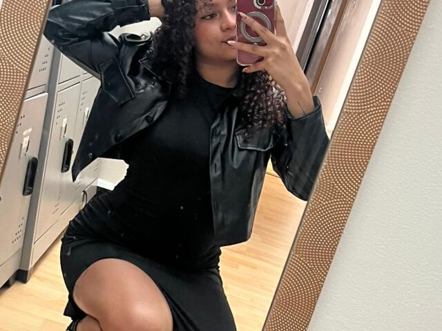 Sofia, passionnée, avec des cheveux bouclés, une veste en cuir noir et une robe noire moulante, montrant ses cuisses dans une photo prise devant un miroir, tenant un téléphone rose, avec un sol en bois et un cadre de miroir texturé..