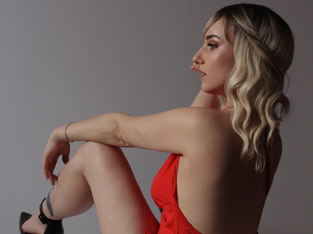 MilenaBaldwin with blonde wavy hair, red dress, and black heels, sitting sideways, showing her back, arms, and legs, with a subtle red light on her right thigh.