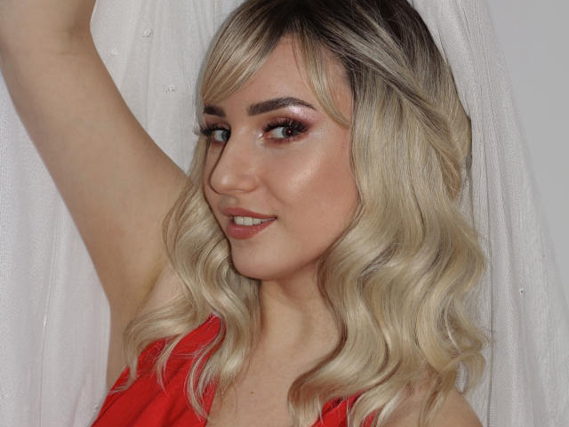 MilenaBaldwin has blonde, wavy hair, red dress, and raised arm. She's smiling with pink eyeshadow, glossy lips, and light makeup. Her tits are small and cute.