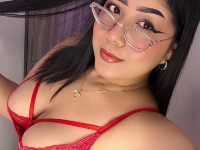 AlexaKiim with glasses, red lace bra showing her tits, black hair, hoop earrings, and a gold necklace, smiling at the camera.