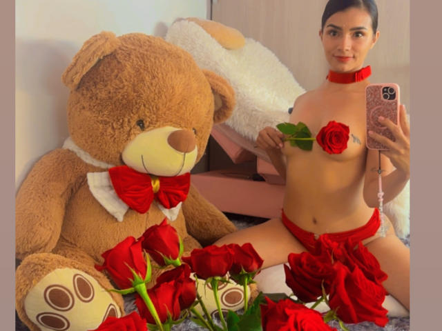 KimmyK com um colar vermelho e um laço vermelho, sem blusa, segurando uma rosa, sentada com um enorme ursinho de pelúcia, cercada por rosas vermelhas. Ela está tirando uma selfie..