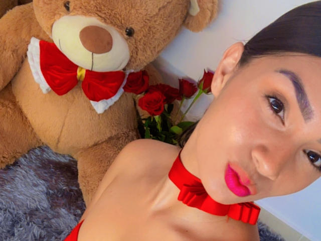 KimmyK, a young woman with fair skin and dark hair, wears a red choker and bright red lipstick. She's positioned in the foreground, her face slightly angled to the right. A large brown teddy bear with a red bowtie and white trim sits behind her, slightly to the left. Red roses with green stems are placed between KimmyK and the teddy bear. The texture of the gray fur beneath them contrasts with KimmyK's smooth skin. The composition is close-up, emphasizing KimmyK's facial features and the teddy bear's plush texture. The colors are bold, with red and brown as the dominant hues.