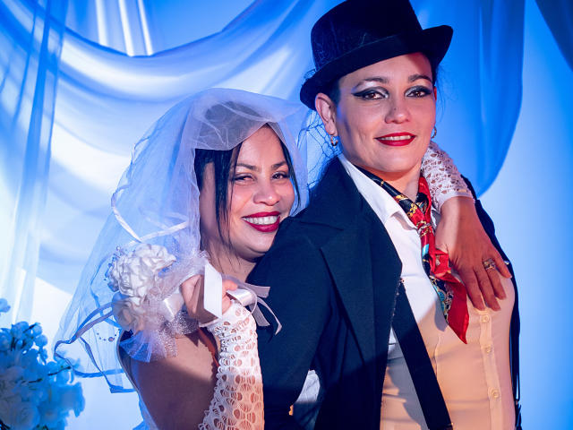 SaharaMK, a bride in a white dress with a veil and floral gloves, stands close to SaharaMK, a woman in a black top hat, black jacket, white shirt, and red tie. SaharaMK on the left has pink lipstick and a smile, while SaharaMK on the right has red lipstick and a confident smile. Blue lighting highlights their faces. SaharaMK's arm is around SaharaMK's shoulder. The image has a playful, formal contrast.