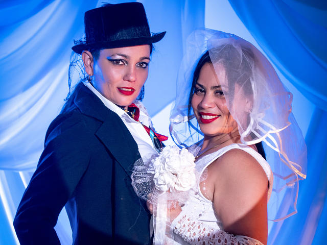 SaharaMK, a woman with red lipstick, wears a white lace dress and a sheer veil. She's next to SaharaMK, a man in a black top hat, black suit, and red tie. Both have white floral brooches. SaharaMK's dress has lace details, and SaharaMK's suit is formal with a white shirt. The man's face is slightly turned, while SaharaMK smiles directly at the camera. The blue-lit background adds a dramatic effect. The image is colorful and highly detailed.