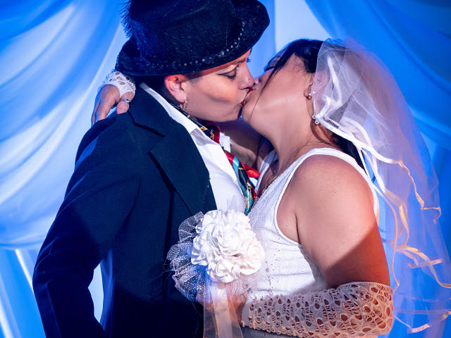 A wedding photo. SaharaMK, a man in a black top hat and dark suit with a colorful tie, kisses SaharaMK, a woman in a white dress and veil, with lace gloves. They're centered, facing each other, with SaharaMK's hand on her back. Soft blue lighting highlights their faces and dresses. White floral accessory on her glove. Close-up.