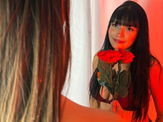 DeseoDuoHot with long black hair, holding red roses in front of her chest, smiling at someone with blonde hair, illuminated by red light. Boobs, roses, red light.