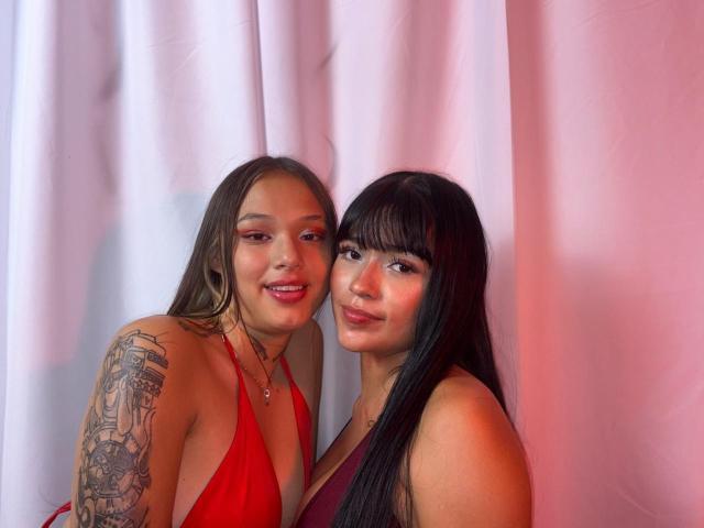 DeseoDuoHot with long brown hair and red dress showing tits, tattoo on arm, smiling next to DeseoDuoHot with black hair and maroon dress, both with makeup.