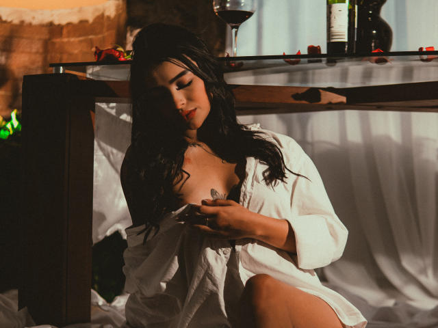 LunaLenox sits in a dimly lit room, wearing a loose white shirt that reveals her tattoo on the right breast. She has long, wet-looking black hair and is looking down at a phone in her right hand. Red rose petals are scattered on a wooden table behind her, and a glass of red wine, a bottle, and a green object are on the table. Soft light casts shadows on her face and body.