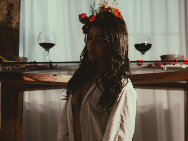 LunaLenox stands in a dimly lit room with soft, warm light. She has long, wavy black hair and a subtle, confident expression. She wears an open white shirt, revealing a dark tattoo on her chest. Two filled wine glasses are on a wooden table behind her, with a floral arrangement and red roses on top. The background is blurred, with sheer white curtains. The image has a moody, intimate atmosphere, with a focus on LunaLenox's relaxed yet slightly mysterious posture.