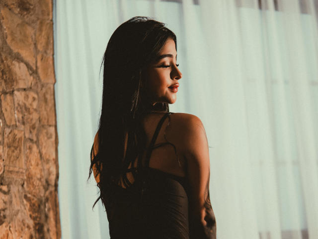 LunaLenox stands with her back to the camera, looking over her shoulder. She has long, dark hair flowing down her back, and her eyes are slightly downcast. She wears a black, sheer-strap top that reveals her shoulders. The light is soft, casting shadows on her face and highlighting her smooth skin. The left side shows a textured stone wall, while the right side is blurred with white curtains. The overall composition is warm and slightly moody.