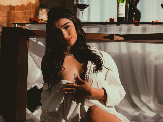 A photograph of LunaLenox with long, dark hair, sitting in soft light. She wears an open white shirt, exposing one small, brown nipple. She holds a wine glass with red wine, her right hand near her chest. Behind her, a wooden table with a glass, wine bottles, and red rose petals. The light casts warm shadows on her skin, highlighting her relaxed, slightly seductive expression. The composition uses warm, natural tones.