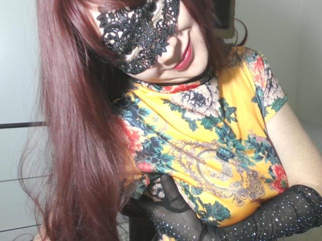 AromaLove with long red hair, wearing a black lace mask, smiles slightly. She's in a yellow floral top with black lace gloves, hands crossed. The mask has intricate lace patterns, and the gloves are sheer with tiny white dots. Her red lipstick contrasts with the lace. The image focuses on her upper body, with a soft light highlighting her face and hands.