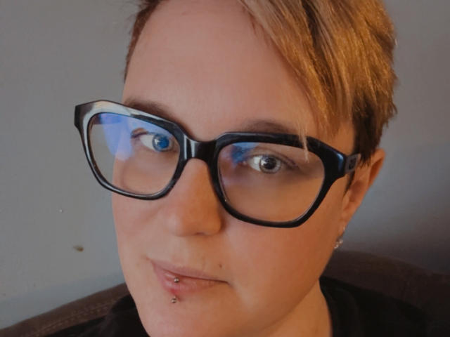 Primer plano de MissLooney con cabello corto y rubio, usando gafas de montura negra y gruesa. Tiene piel clara, ojos azules y un pequeño piercing plateado en el labio inferior. Está mirando directamente a la cámara con una sonrisa leve y segura. La iluminación es suave, resaltando su piel clara y el maquillaje sutil. Lleva una camisa negra, y el fondo es una pared simple de color claro. La imagen es nítida con un tono de color natural y ligeramente cálido..