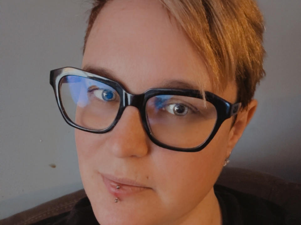 Primer plano de MissLooney con cabello corto y rubio, usando gafas de montura negra y gruesa. Tiene piel clara, ojos azules y un pequeño piercing plateado en el labio inferior. Está mirando directamente a la cámara con una sonrisa leve y segura. La iluminación es suave, resaltando su piel clara y el maquillaje sutil. Lleva una camisa negra, y el fondo es una pared simple de color claro. La imagen es nítida con un tono de color natural y ligeramente cálido..