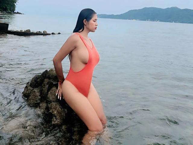 Sensualbunny, une femme aux cheveux foncés, agenouillée sur une plage, porte une combinaison rouge en dentelle et montre ses seins et ses fesses. Une falaise rocheuse et un mur de pierre se trouvent en arrière-plan..
