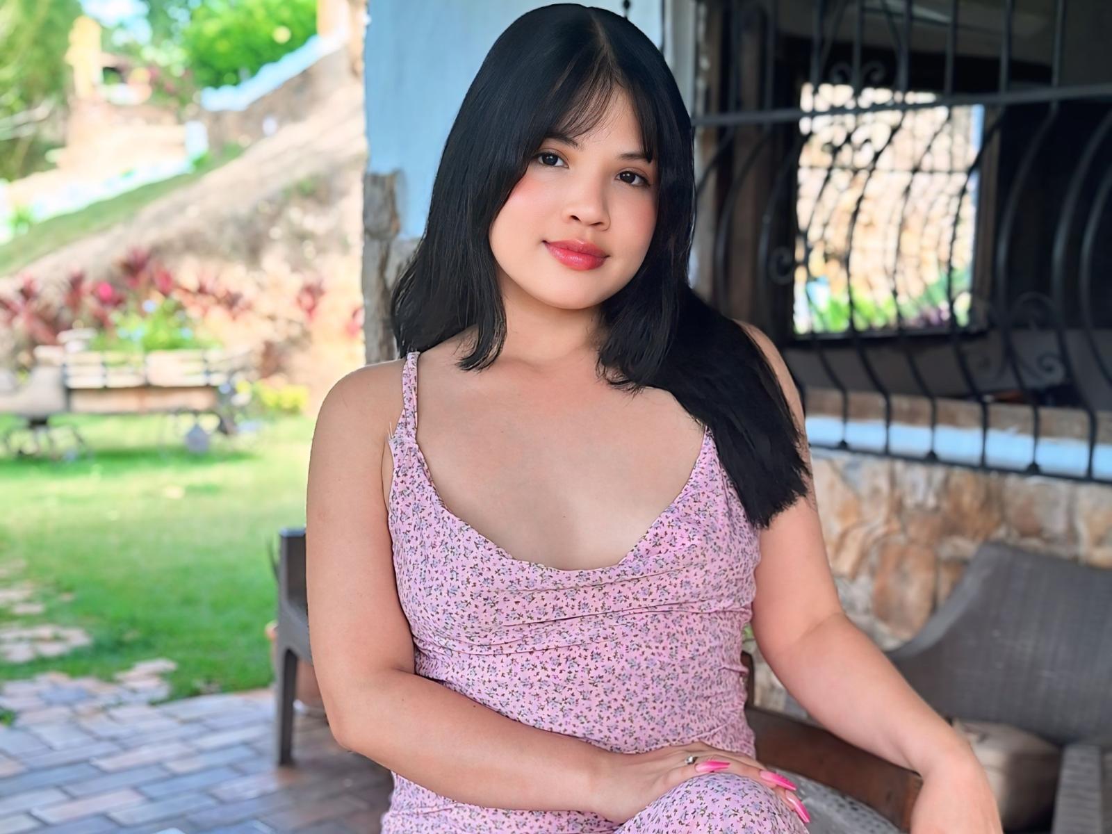 Photograph of MissLilyh, a young Asian woman with straight black hair and light skin. She wears a pink floral dress with a deep V-neck, exposing part of her chest. She sits on a wooden chair with one hand on her lap. Her facial expression is neutral, with a slight smile. The background is blurred, showing a green lawn and a stone wall. Her nails are painted pink. The image is brightly lit with natural light.