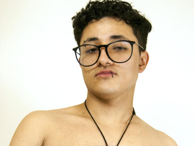 AdoniBaskerville has curly black hair, large black glasses, and a small lip piercing. He's shirtless with a black necklace, and has a slightly pouty expression.