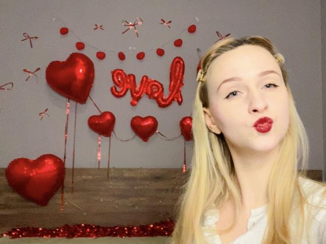 FlyFreedom has long blonde hair, a white shirt, and is pouting with red lipstick. Red heart balloons and a red "val" sign are in the background.