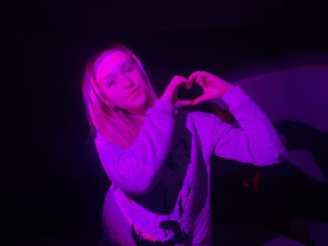 FlyFreedom with blonde hair, pink neon light, white hoodie, forming a heart shape with hands, looking slightly to the side, sitting in a dark, purple-lit environment.