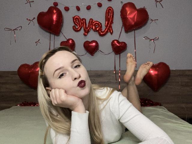 FlyFreedom with blonde hair, white long-sleeve shirt, and dark lipstick, pouting, lying on bed with feet up, surrounded by red heart balloons and "swol" in background.