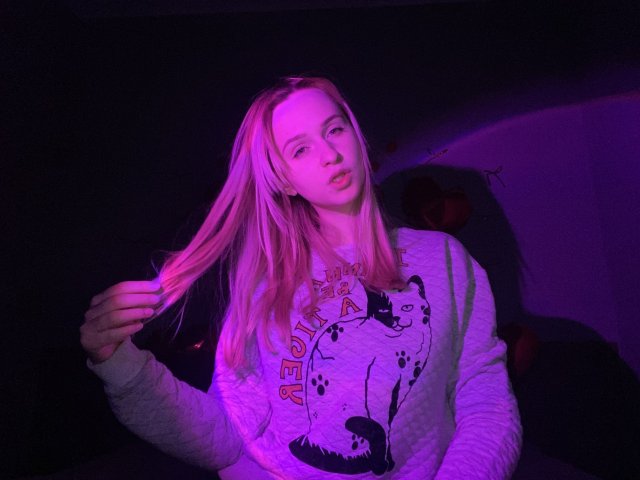 FlyFreedom with long blonde hair, pink-lit face, white graphic sweatshirt, holding hair, purple and pink lighting, slightly open mouth, looking at camera, dark background.