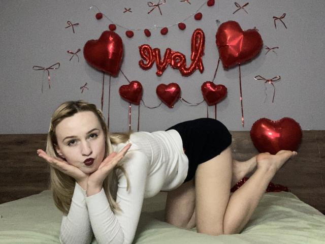FlyFreedom, blonde with fair skin, on bed, white long-sleeve, black shorts, knees up, hands on face, looking at camera, red heart balloons, red "val" letters, red ribbons.
