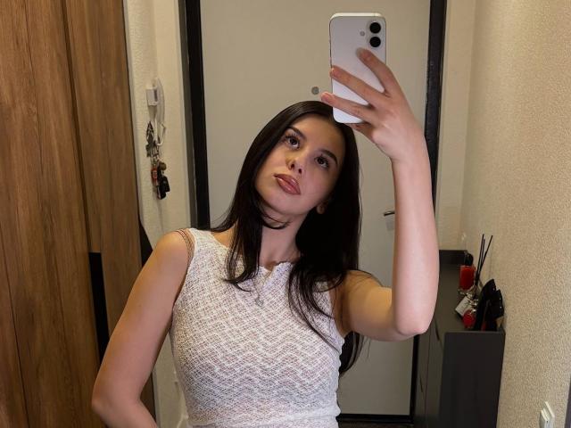 DanyellEden with long black hair, white textured sleeveless top, holding phone, taking mirror selfie, showing small tits, pale skin, red lipstick, wooden door, and white wall in background.