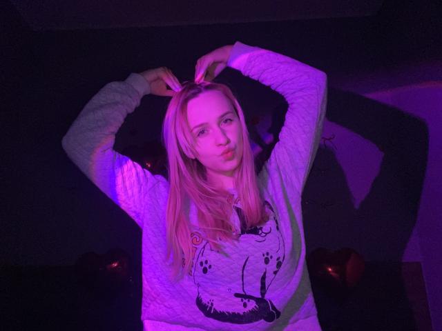 FlyFreedom with blonde hair, pouty lips, and purple-lit face, wearing a white hoodie with paw prints, arms raised, hands in hair, looking at camera.