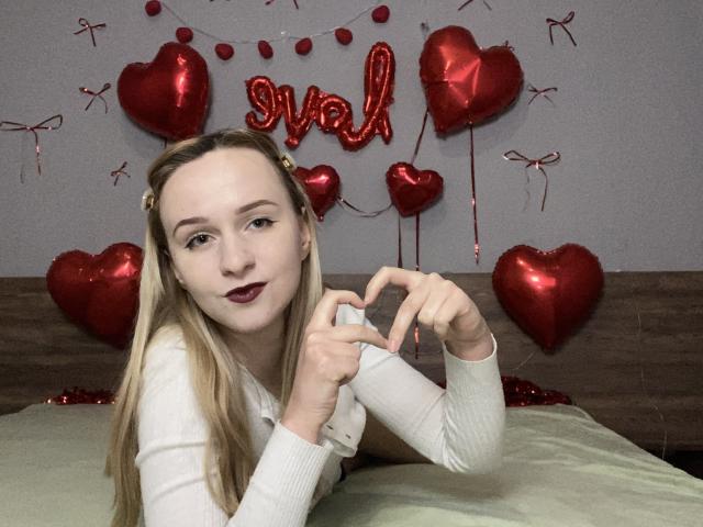 FlyFreedom with blonde hair, wearing a white top, making a heart shape with her hands, red balloon hearts in the background, and dark red lipstick, lying on a bed.